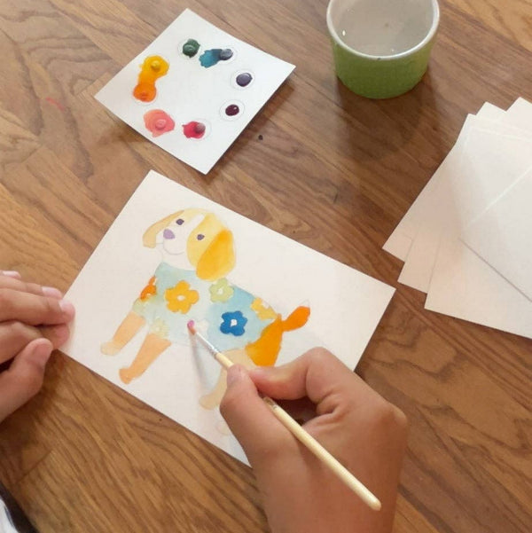 Kid painting a dog postcard with watercolors on a wood table.