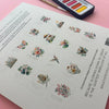 The back cover of a watercolor art workbook showing examples of the nature-themed designs included in the book. There is a watercolor palette and paintbrush next to the book resting on a pink background.