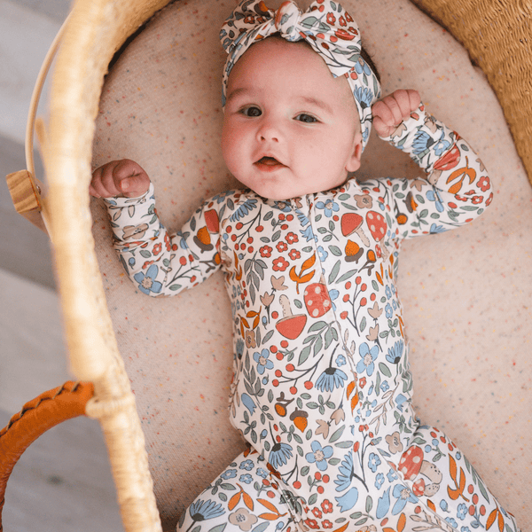 A baby lying on their back inside a woven bassinet, wearing a long-sleeve floral print romper and a matching headband.
