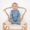A baby sitting on a wooden chair wearing a blue footed zip-up sleeper with a dog print, photographed against a plain white background.