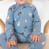 A baby wearing a light blue long-sleeve zip-up sleeper with an all-over print of different dog breeds, sitting on a woven chair.