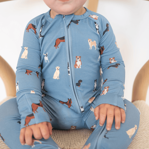 A baby wearing a light blue long-sleeve zip-up sleeper with an all-over print of different dog breeds, sitting on a woven chair.