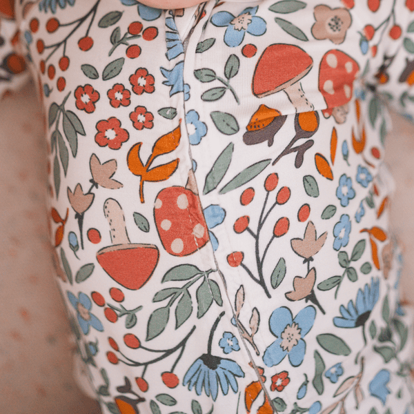 Close-up of a baby's chest wearing a white long-sleeve romper with a colorful print of red-capped mushrooms, blue flowers, green leaves, and orange berries.