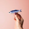 Hand holding a small blue and white fish-shaped hair claw against a pink background