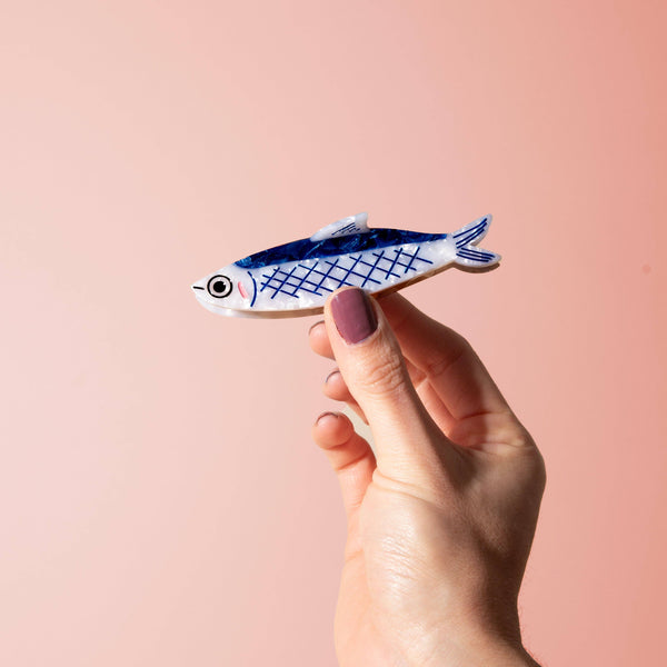 Hand holding a small blue and white fish-shaped hair claw against a pink background
