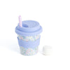 A kid-sized reusable travel tumbler cup with a pale blue silicone lid and lavender straw, featuring a floral patterned body and a pale blue silicone sleeve. A small white marshmallow is placed beside it on a clean white background.