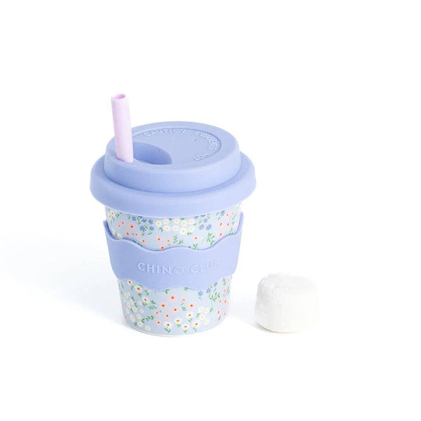 A kid-sized reusable travel tumbler cup with a pale blue silicone lid and lavender straw, featuring a floral patterned body and a pale blue silicone sleeve. A small white marshmallow is placed beside it on a clean white background.