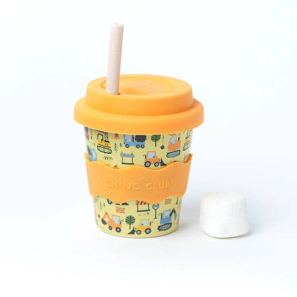 Kid sized travel cup in yellow printed with construction themed motifs, golden yellow silicone wavy grip, lid, and white straw. A marshmallow sits next to the cup against a white backdrop.