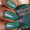 Close-up of green glittery nail polish on a hand with a bottle labeled 'Cirque Colors'.
