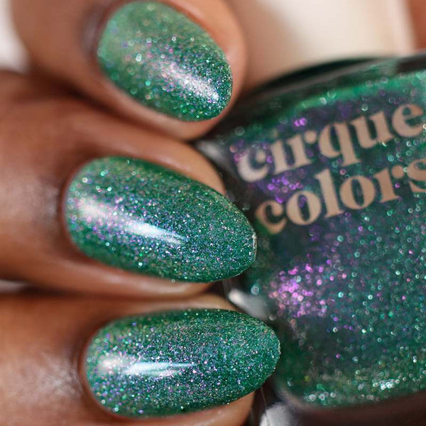 Close-up of green glittery nail polish on a hand with a bottle labeled 'Cirque Colors'.