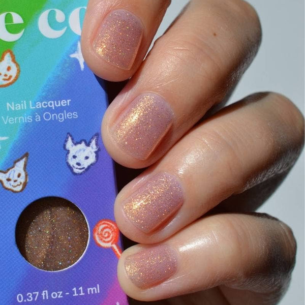 A close-up of a hand with glossy nude pink nail polish featuring a subtle gold shimmer, holding a colorful product box labeled 'cirque colors Nail Lacquer' with a circular window showing the peachy pink glitter polish inside.