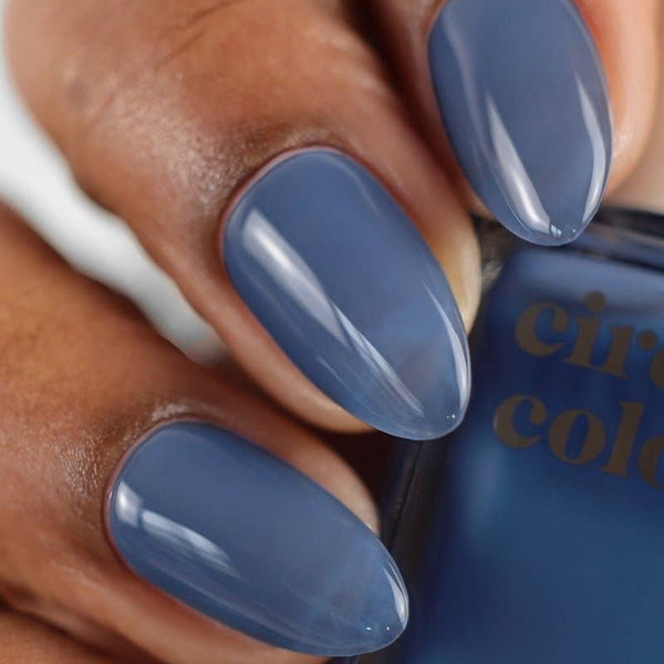 Close-up of a hand with almond-shaped nails painted in a dusty slate blue shade, holding a bottle of Circolor nail polish.