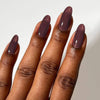 A close-up of a hand with long, almond-shaped nails painted in a glossy, muted mahogany shade against a plain light background.