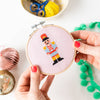Cross-stitched nutcracker design held in an embroidery hoop with crafting materials in the background.