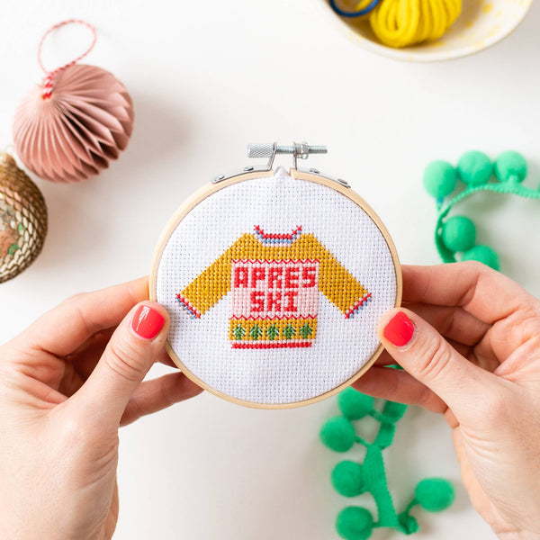Hands holding a wooden embroidery hoop with a cross-stitch design of a yellow sweater that says 'APRES ski' in red letters, surrounded by festive craft props on a white background.
