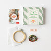 A cotton cross-stitch kit laid out on a white background, including a striped product box labeled 'Cotton Clara Small Shop Cross Stitch Kit,' a wooden embroidery hoop, white fabric, a needle, thread, and a printed instruction card.