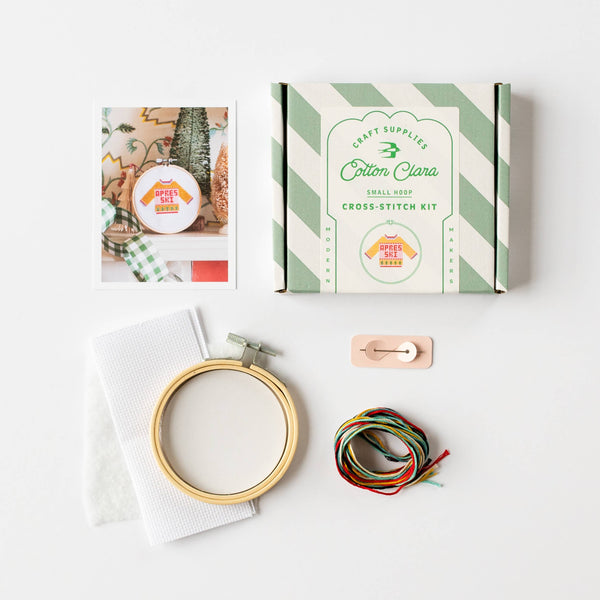 A cotton cross-stitch kit laid out on a white background, including a striped product box labeled 'Cotton Clara Small Shop Cross Stitch Kit,' a wooden embroidery hoop, white fabric, a needle, thread, and a printed instruction card.