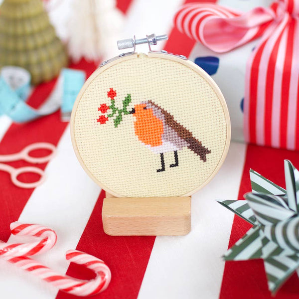 Cross-stitched robin design in a hoop on a red fabric background with Christmas decorations.