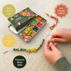 A children's forest-themed beaded bracelet craft kit shown open in a metal tin with multiple color and shape beads, alongside finished beaded strands and a pair of hands actively making a bead necklace. Text callouts around the scene say '100% Plastic Free,' 'Eco Friendly Craft Kit,' 'All Inclusive Set,' and 'Enhances Motor Skills.