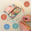 A bracelet beading craft kit shown on a light beige fabric background, with an open teal tin of colorful beads, a pink branded box, and a beaded cord being threaded by hands. Text callouts read 'Eco Friendly Craft Kit,' '100% Plastic Free,' 'All Inclusive Set,' and 'Enhances Motor Skills.