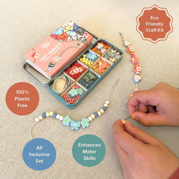 A bracelet beading craft kit shown on a light beige fabric background, with an open teal tin of colorful beads, a pink branded box, and a beaded cord being threaded by hands. Text callouts read 'Eco Friendly Craft Kit,' '100% Plastic Free,' 'All Inclusive Set,' and 'Enhances Motor Skills.