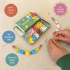 A children's beaded bracelet kit shown on a light beige surface with a kid's hands assembling beaded bracelets. The set includes a green Wildflower branded tin box, colorful round beads, flower-shaped beads, and leaf shaped beads.