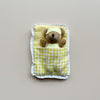A small plush teddy bear wearing a yellow polka-dot sleep mask and matching pajamas, tucked inside a yellow-and-white gingham sleeping bag with a white lace trim, set against a plain beige background.