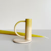 A taper candle holder with curved handle. It is enameled with a pink to yellow 'sunset' gradient. There is a yellow taper candle placed behind the holder against a neutral background.
