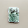 A small gray plush elephant toy wearing striped pajamas and a matching sleep mask, tucked inside a soft fabric sleeping bag with a green plaid lining and white lace trim, shown against a plain beige background.