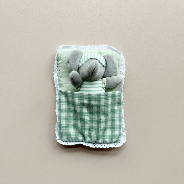 A small gray plush elephant toy wearing striped pajamas and a matching sleep mask, tucked inside a soft fabric sleeping bag with a green plaid lining and white lace trim, shown against a plain beige background.
