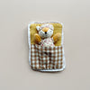 A small plush fox toy wearing floral pajamas and a matching sleep mask, tucked inside a fabric brown plaid sleeping bag and white lace trim, photographed against a plain light beige background.
