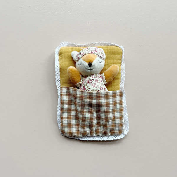 A small plush fox toy wearing floral pajamas and a matching sleep mask, tucked inside a fabric brown plaid sleeping bag and white lace trim, photographed against a plain light beige background.