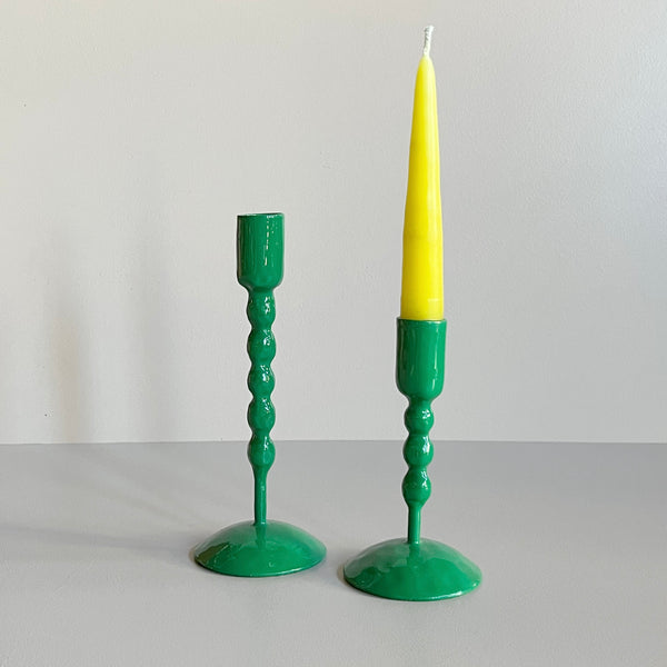 A pair of tall and short bright green enameled cast iron taper candle holders with bubble textured shafts, the short one sporting a yellow taper candle, against a neutral backdrop.