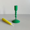 A short bright green enameled iron taper candle holder with a yellow taper candle resting in front against a neutral backdrop.