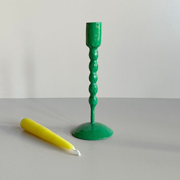 A tall bright green enameled iron taper candle holder with a yellow taper candle resting in front against a neutral backdrop.