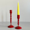 A pair of tall and short bright red enameled cast iron taper candle holders with bubble textured shafts, the short one sporting a yellow taper candle, against a neutral backdrop.