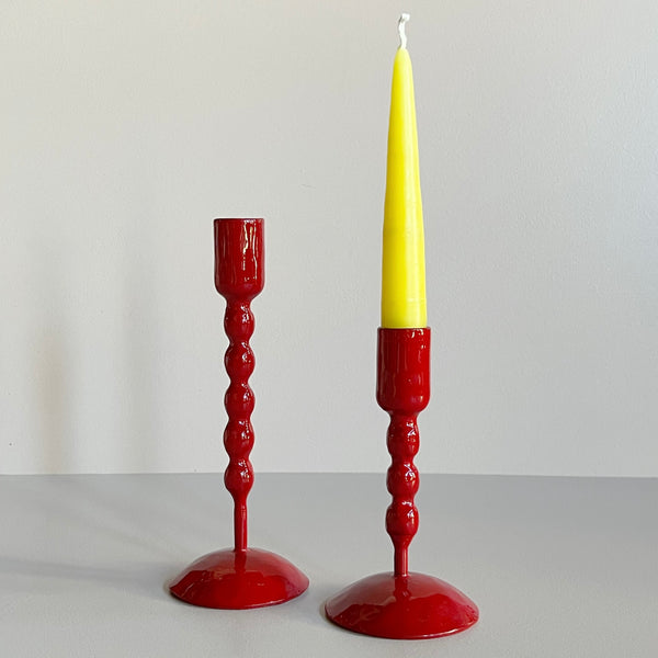 A pair of tall and short bright red enameled cast iron taper candle holders with bubble textured shafts, the short one sporting a yellow taper candle, against a neutral backdrop.