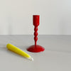 A short bright red enameled iron taper candle holder with a bright yellow candle resting in front of it on a neutral backdrop.