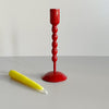 A tall bright red enameled iron taper candle holder with a bright yellow candle resting in front of it on a neutral backdrop.