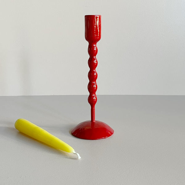 A tall bright red enameled iron taper candle holder with a bright yellow candle resting in front of it on a neutral backdrop.