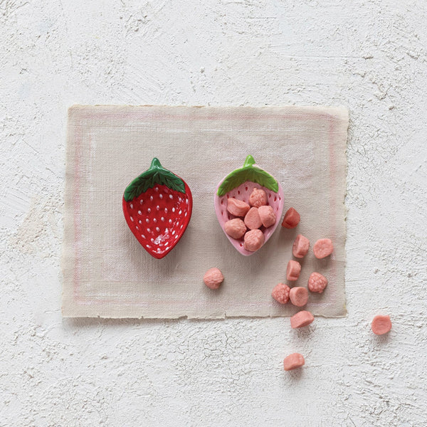 One empty red strawberry shaped dish and one pink strawberry shaped dish overflowing with pink gummy vitamins on a neutral tabletop.