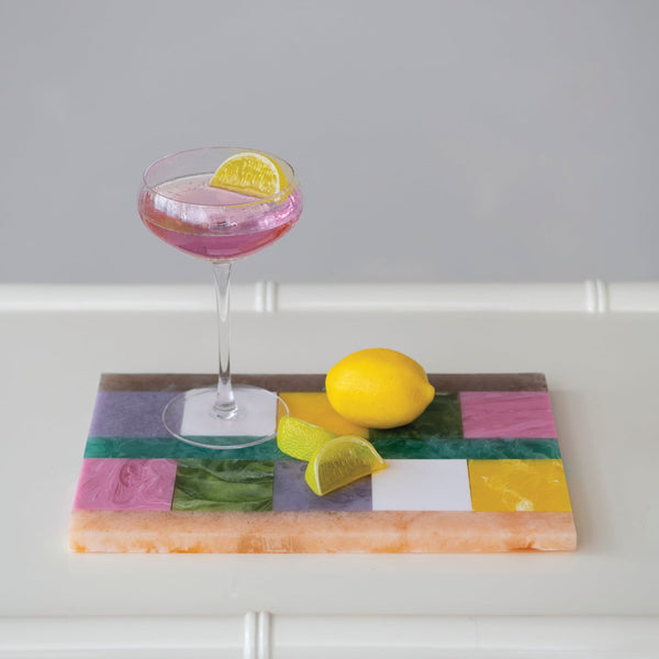 A rectangular marbled resin serving board with a colorful patchwork design, styled with a pink cocktail in a coupe glass, a whole lemon, and a lime wedge on a white table against a light gray background.