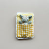A small gray plush mouse wearing a floral sleep mask and pajamas, tucked inside a yellow-and-white checkered quilted sleeping bag with a white lace trim, set against a plain light gray background.