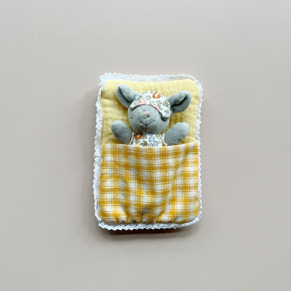 A small gray plush mouse wearing a floral sleep mask and pajamas, tucked inside a yellow-and-white checkered quilted sleeping bag with a white lace trim, set against a plain light gray background.