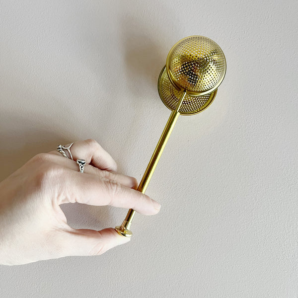 A hand plunging open a gold toned stainless steel tea infuser.