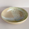 A large low serving bowl in a soft tan featuring a watercolor reactive speckled glaze in a mix of yellow, green and blue against a neutral background.