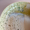 A detailed view large low serving bowl in a soft tan featuring a watercolor reactive speckled glaze in a mix of yellow, green and blue against a neutral background.