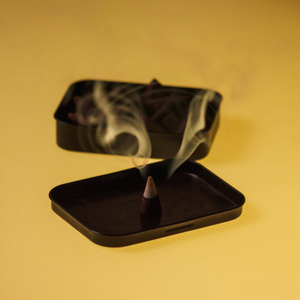 A black metal incense burner set with a tray and lid, shown with a smoking incense cone in the foreground and more incense cones inside the lid in the background, set against a warm yellow background.