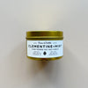 A gold metal tin candle by 'Fern & Nettle' labeled Clementine + Mint against a neutral background. 