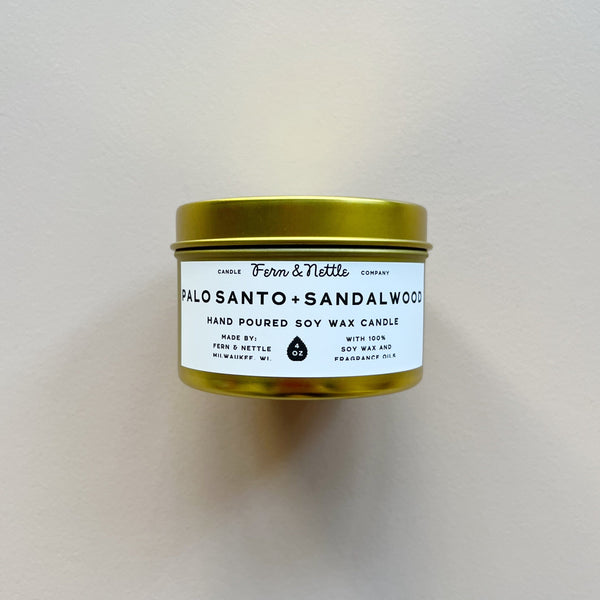 A gold metal candle tin by 'Fern & Nettle' labeled Palo Santo + Sandalwood against a neutral background.